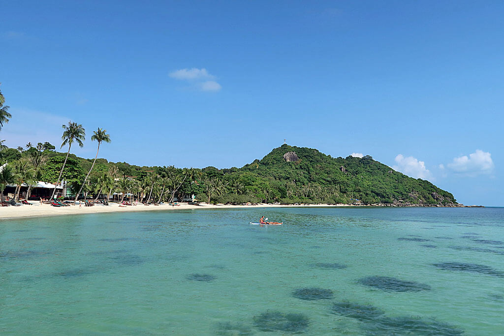 Ein Strand mit blauem Meer, ein Kajak und Hügel mit Palmen und Felsen