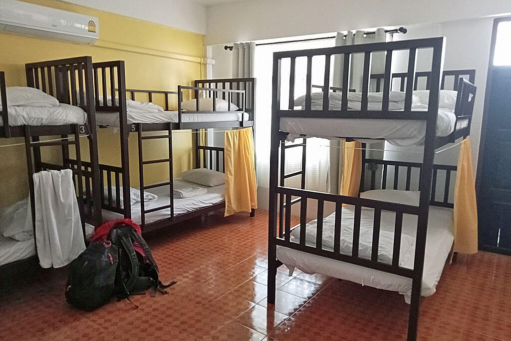 Ein Mehrbettzimmer mit drei Stockbetten, rot gefliester Boden, weiße Bettwäsche und ein Rucksack auf dem Boden