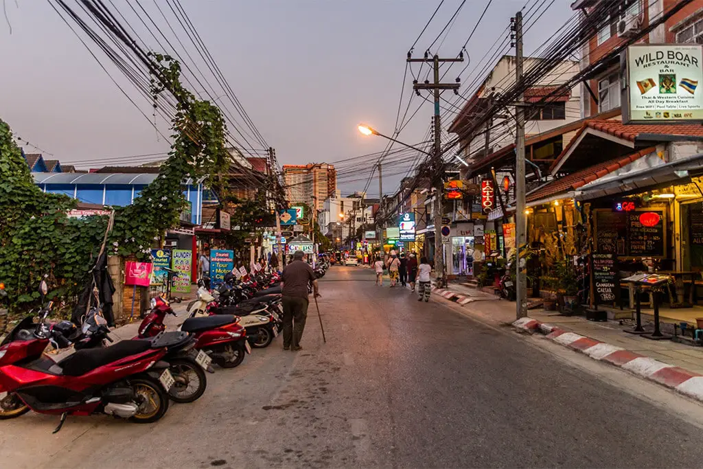 Straße in Chiang Mai im Abendlicht mit Mann mit Stock und geparkten Mopeds