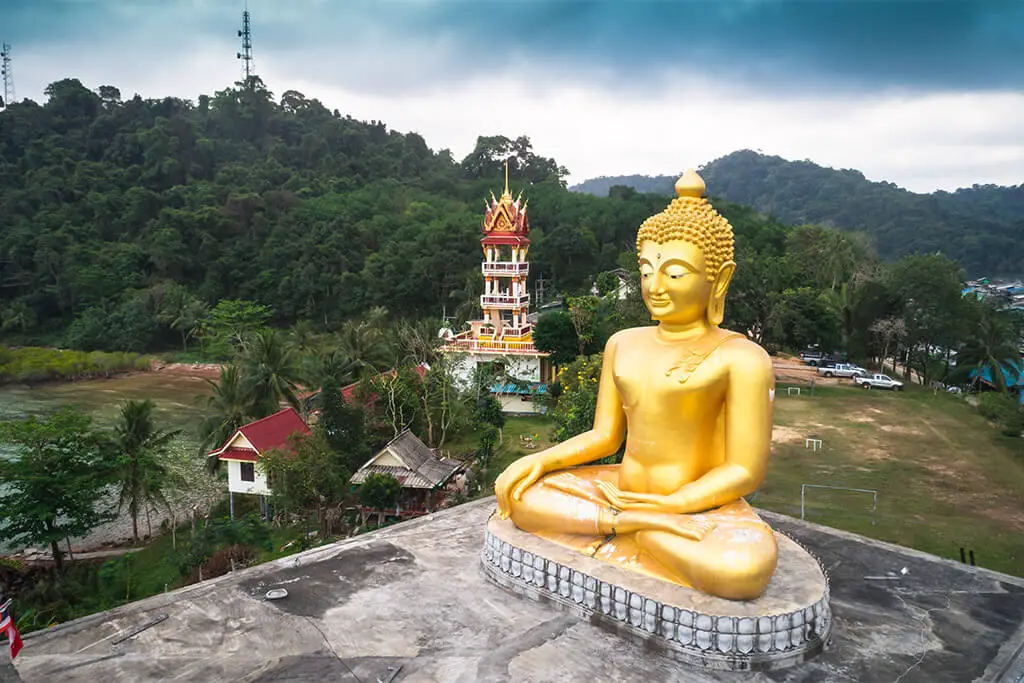 Golden Buddha Staue vor einem Tempelturm umgeben von grüner Landschaft