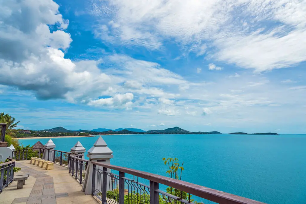 Ein Weg mit Geländer führt zu einem Ausblick auf das blaue Meer und über die hügelige Insel