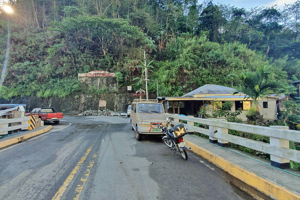 Eine breite Straße mit Fahrzeugen, dahinter beginnt der Regenwald