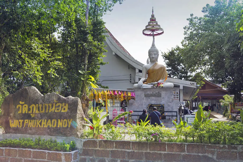Eine kleine Tempelanlage mit einem weißen, sitzenden Buddha, einem Schriftzug im Felsen und betenden Menschen