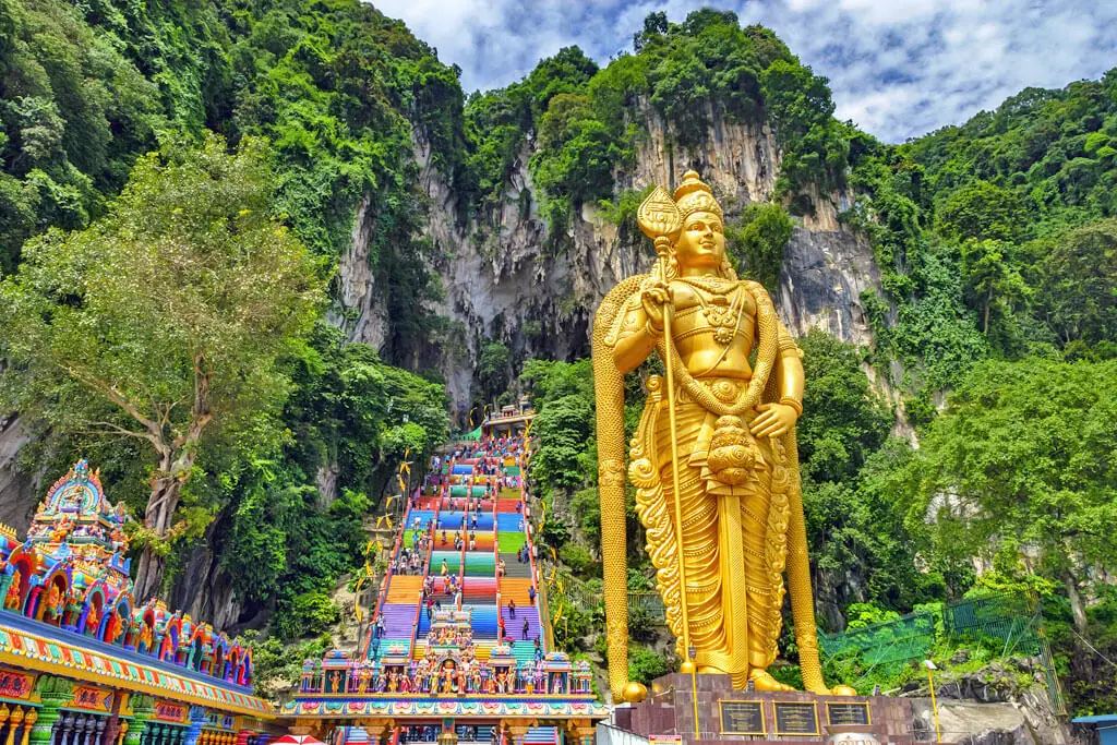 Eine lange Treppe mit bunten Stufen führt einen Berg hinauf, davor thront die goldene Statue der Gottheit Murugan