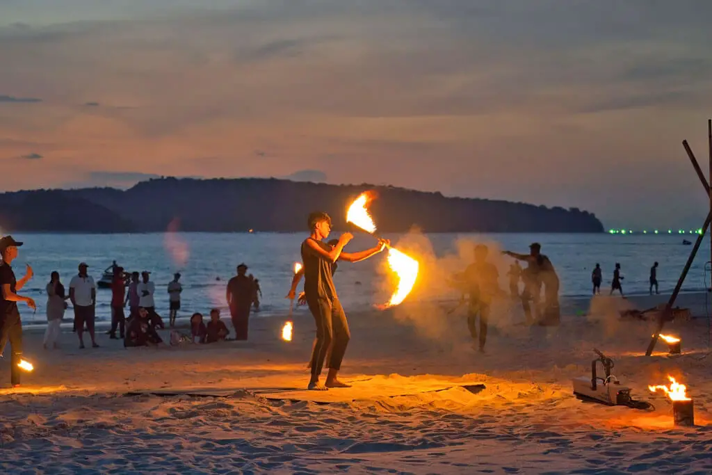 Junge Männer bei ihrer Feuershow am Strand bei Sonnenuntergang, dahinter viele Menschen