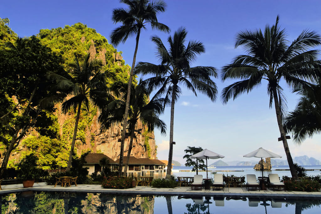 Hotelpool mit Sonnenliegen und Sonnenschirmen, Palmen und dem Meer und Felsen im Hintergrund