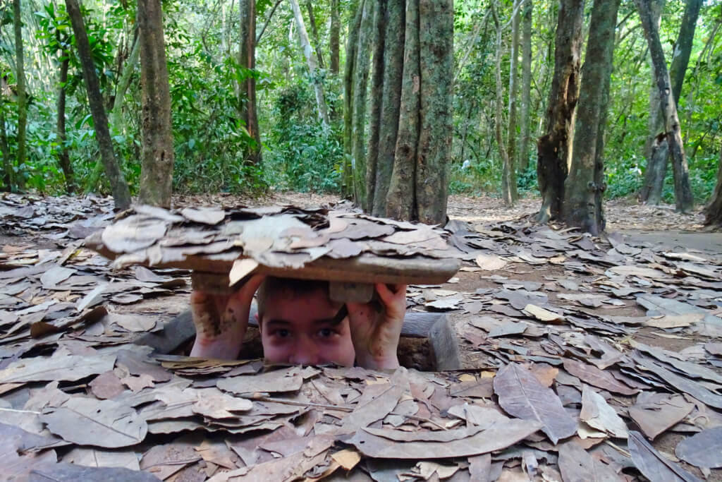 Der Kopf eines jungen Mannes schaut im Wald aus einem Loch im Boden, das mit einem Deckel verschlossen ist, den der junge Mann über dem Kopf hält.