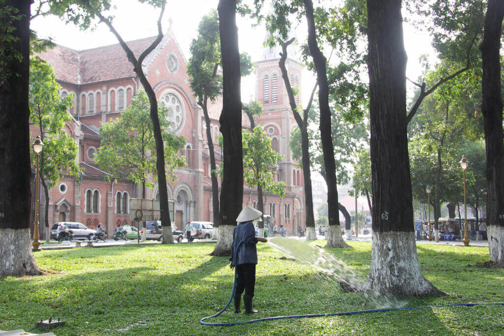 Eine vietnamesische Frau mit konischem Hut wässert den Rasen in einem Park in Ho-Chi-Minh-Stadt, im Hintergrund steht die Notre-Dame-Kathdrale.