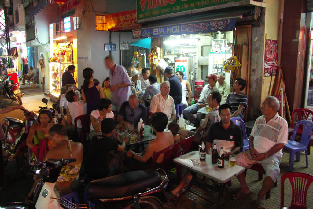 Nachtaufnahme einer belebten improvisierten Bar mit kleinen Tischen und Plastikstühlen auf einem Bürgersteig in Ho-Chi-Minh-Stadt. 