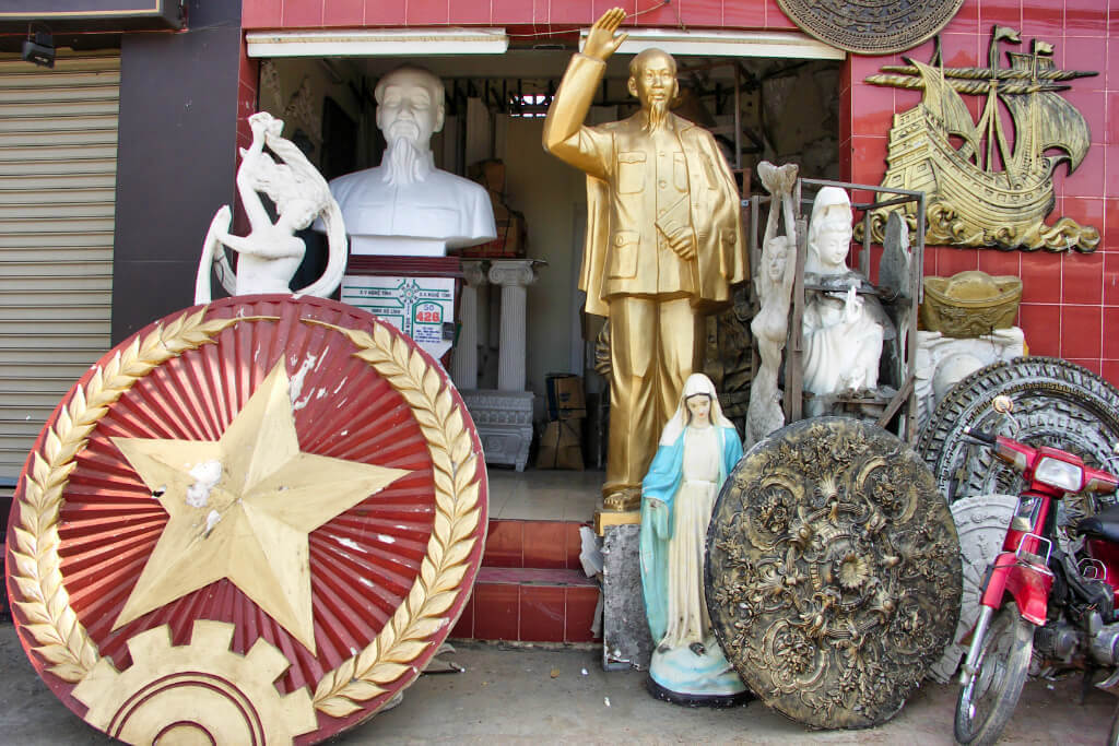 Auf einem Bürgersteig vor einem kleinen Geschäft stehen verschiedene Statuen und Reliefs, darunter eine goldene, lebensgroße Statue von Ho Chi Minh.