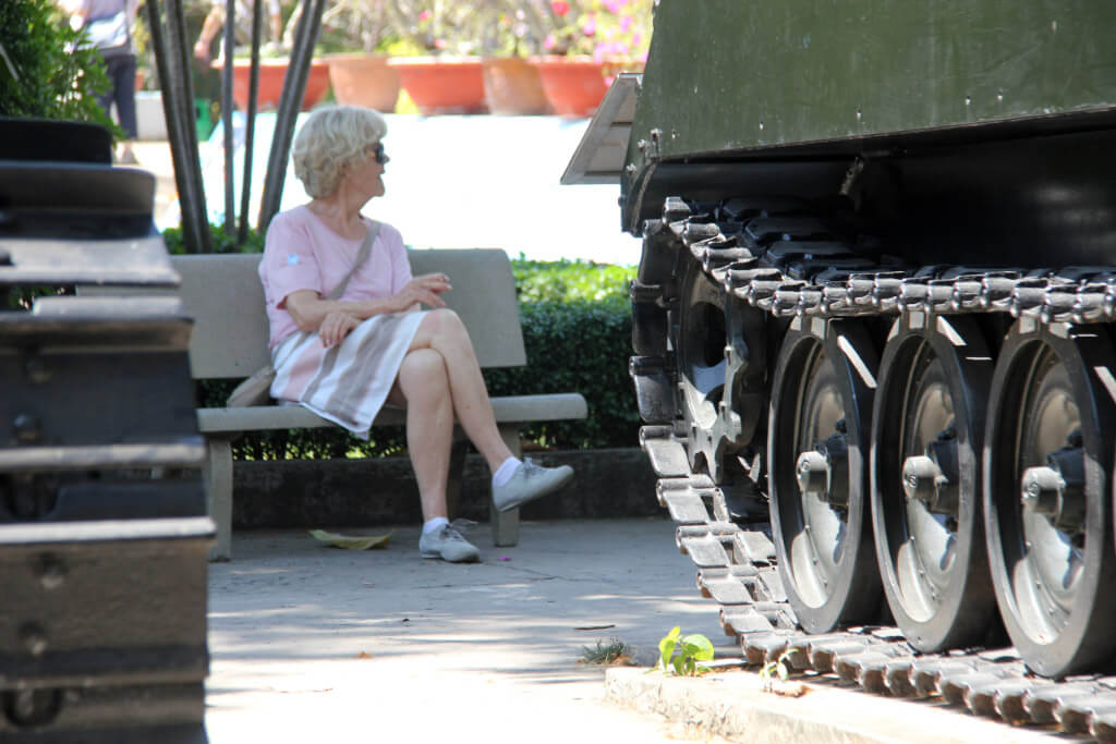 Eine ältere Frau sitz auf einer Bank; davor stehen zwei Panzer, von denen nur Teile des Kettenantriebs zu sehen sind.