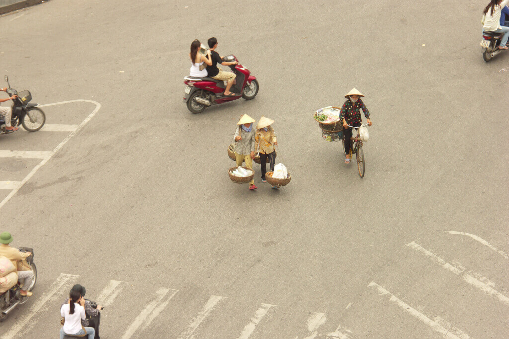 Hanoi, Straßenkreuzung am Hoan-Kiem-See. (c) Mark Markand Straßenkreuzung von oben gesehen, mit wenigen Motorrädern, einem Fahrrad und zwei Frauen mit konischen Hüten und Tragestange