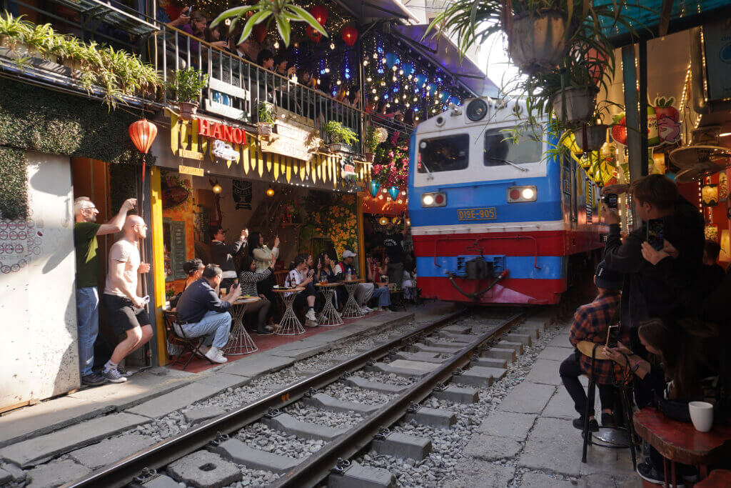 Train Street, Hanoi. (c) Andrea Markand Ein Zug fährt durch eine sehr enge, mit Gleisen versehene Gasse, an deren Rand Touristen in einem Café sitzen und Fotos machen.