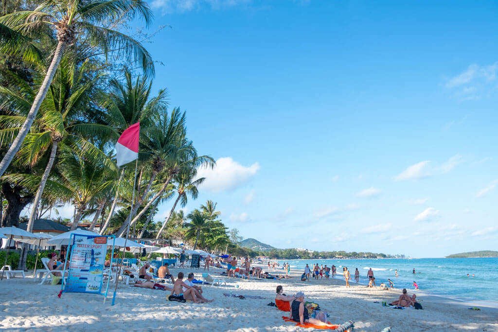 Menschen sitzen oder spazieren am Strand, Schild mit Wassersport-Angeboten und rot-weiße Fahne im Vordergrund, im Hintergrund Palmen und grüne Hügel