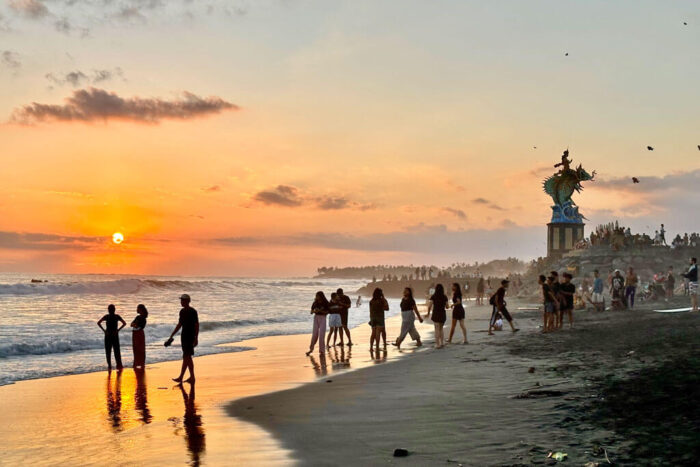 Ein Strand bei Sonnenuntergang mit Menschen, einer großen Skulptur und der untergehenden Sonne über dem Meer