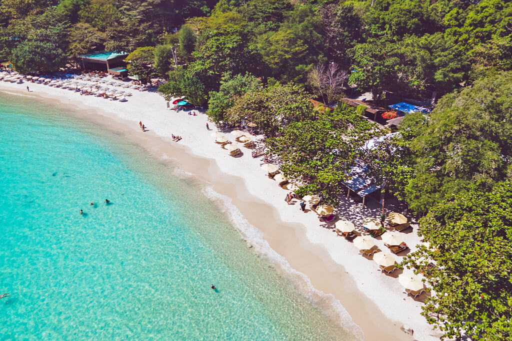 Luftaufnahme auf einen Strandabschnitt mit Sonnenschirmen, Palmen und wenigen Hotels