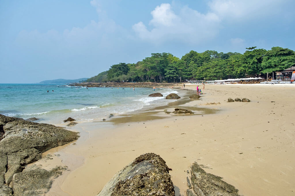 Ein breiter Sandstrand mit großen Felsen und einigen Badegästen im Wasser