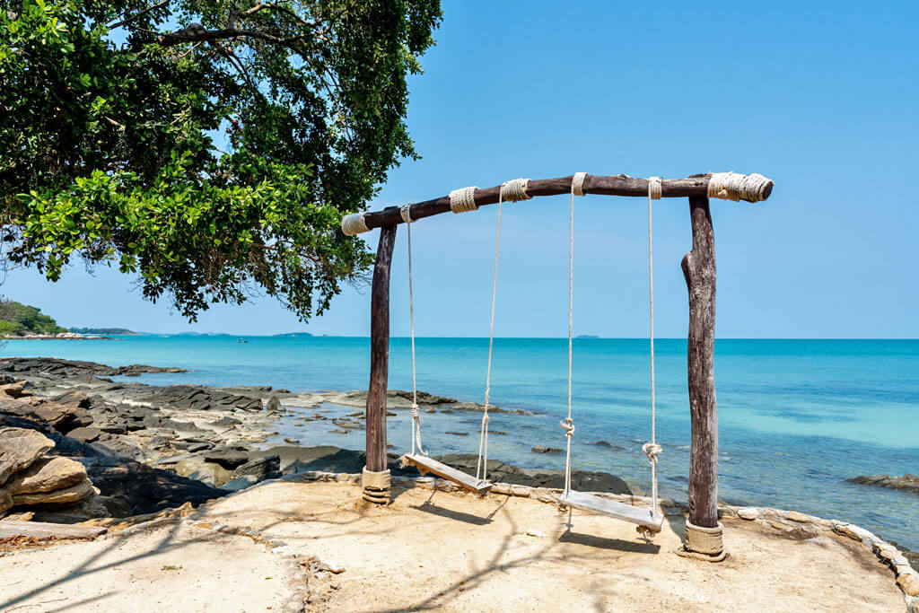 Zwei Holzschaukeln an einem felsigen Strand mit einem Baum
