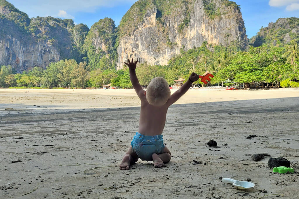 Ein Baby in Windeln an einem Strand, dahinter Felsen