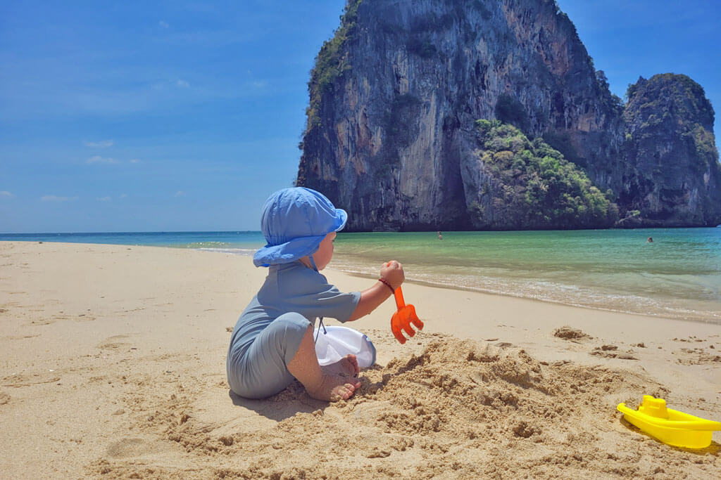 Ein Baby im UV-Anzug spielt am Strand mit Sandelsachen, im Hintergrund ein Felsen im Meer