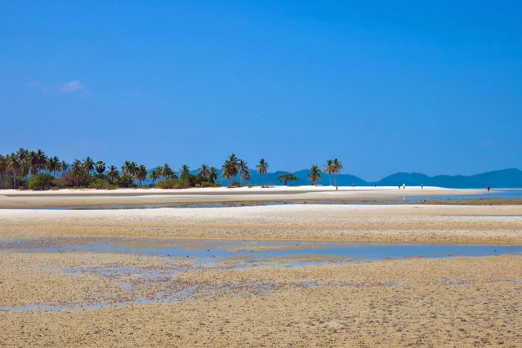Eine breite Sandbank bei Ebbe, im Hintergrund Palmen und Inseln am Horizont