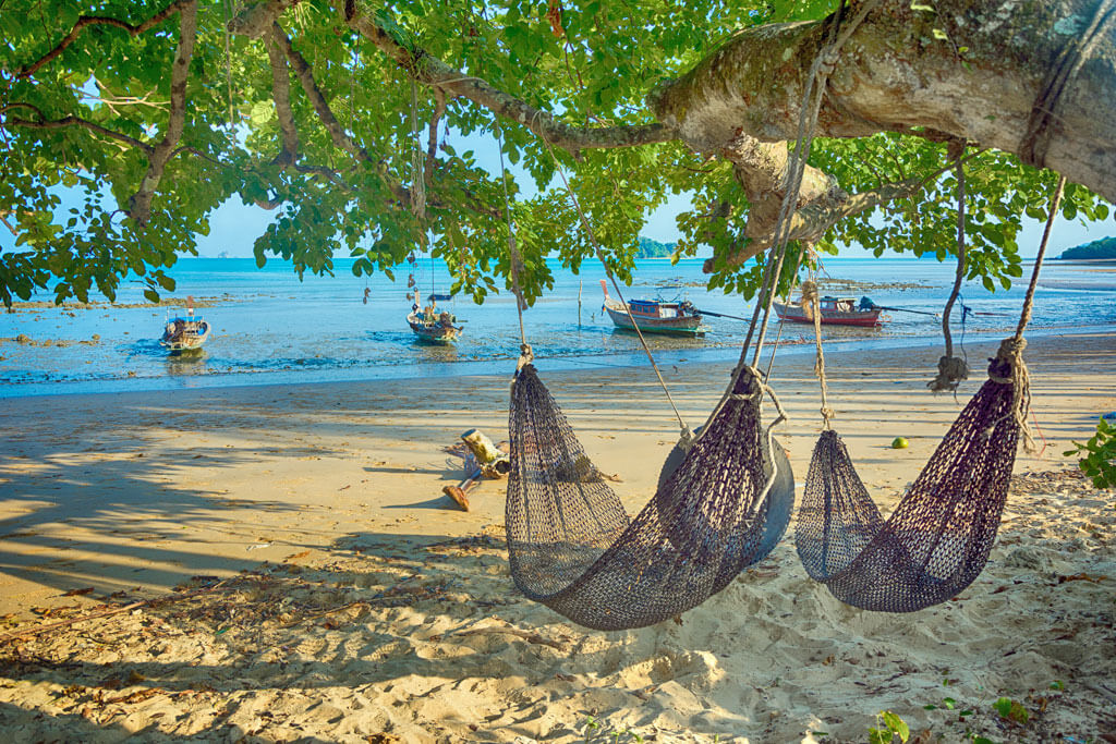 Zwei Hängematten hängen an Bäumen direkt an einem Strand bei Ebbe, Longtailboote liegen auf dem Sand