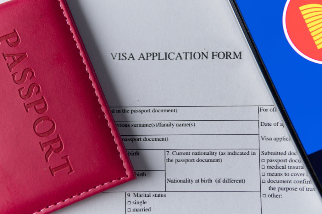 Visa Application Form auf Englisch in Papierform, auf dem Dokument liegt ein Reisepass in einer Hülle mit der Aufschrift "Passport" und einem Handy, auf dem zur Hälfte die ASEAN-Flagge zu sehen ist