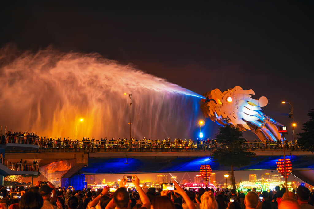 Die bunt beleuchtete Drachenbrücke in Danang speit bei Nacht Wasser, während viele Zuschauer mit ihren Mobiltelefonen davon Fotos machen