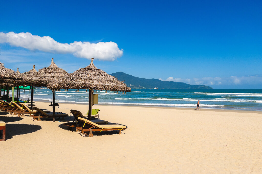 Da Nang, My Khe Strand, mit strohgedeckten Sonnenschirmen, Strandliegen und der Son-Tra-Halbinsel im Hintergrund