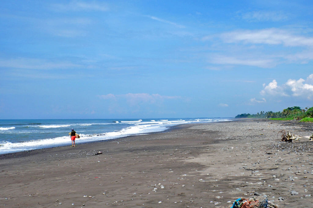 Surfer am schwarzen Sandstrand von Medewi auf Bali vor blauem Himmel