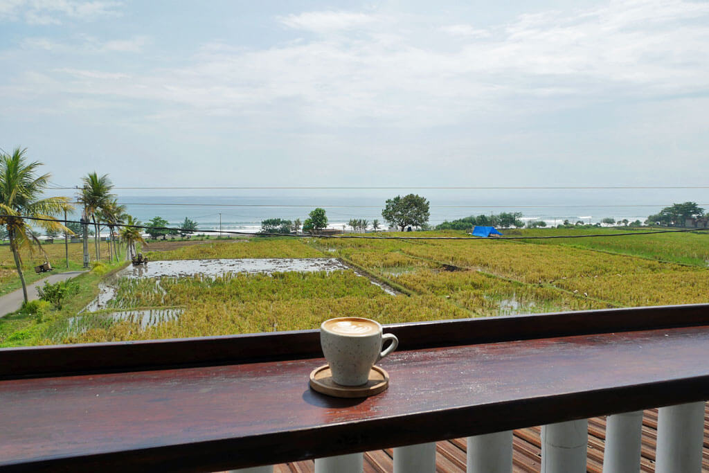 Kaffeetasse auf einer hölzernen Brüstung mit Blick über Reisfelder und das Meer von Medewi auf Bali