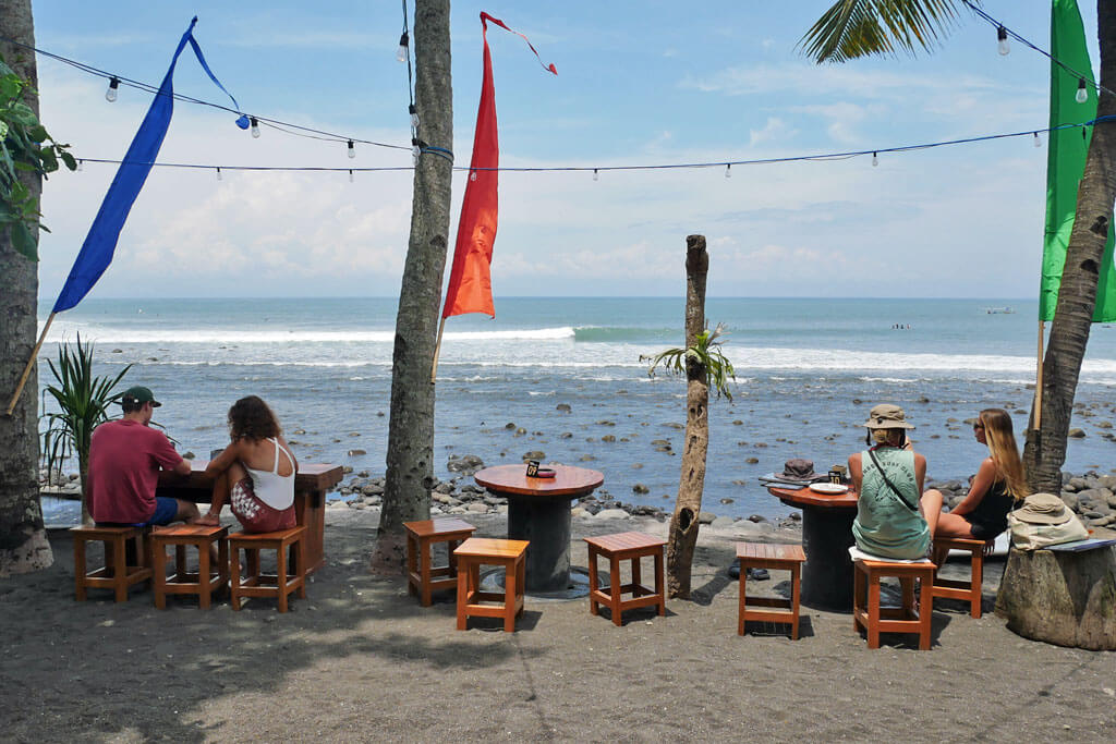 Touristen sitzen auf Holzhockern mit direktem Blick auf das Meer von Medewi Beach bei blauem Himmel und Sonnenschein