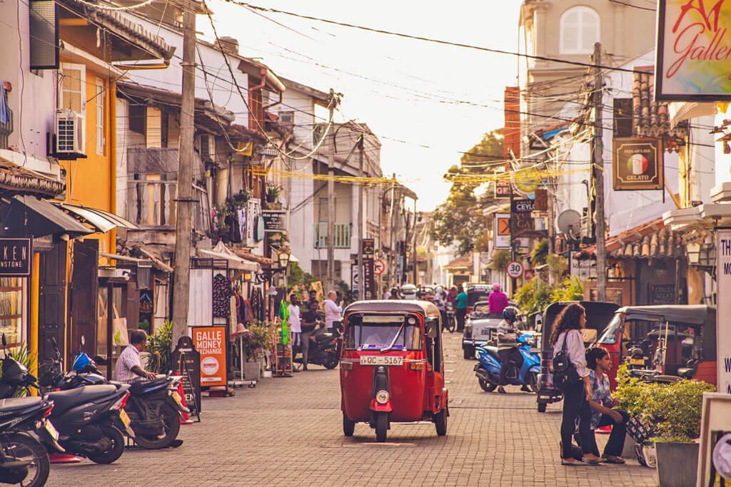 Eine lebhafte Straße mit Tuk Tuk, Häusern, Stromkabeln und Rollern in Abendstimmung
