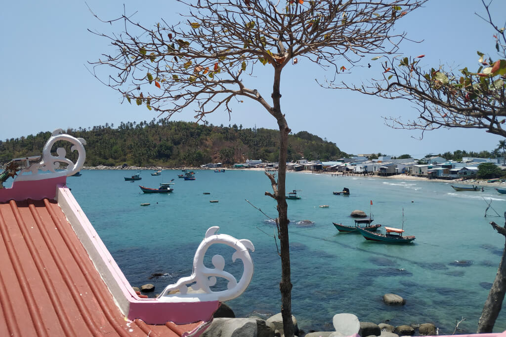 Blick über eine Bucht mit kleinen Booten auf ein Fischerdorf auf Hon Son; im Vordergrund im Anschnitt ein Tempeldach mit roten Ziegeln