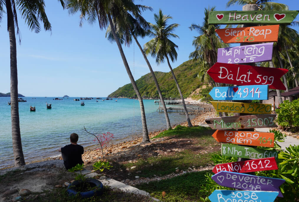 Wegweiser an einem kiesigen Strandabschnitt auf Nam Du in Vietnam, ein junger Mann in Rückansicht blickt auf die Bucht